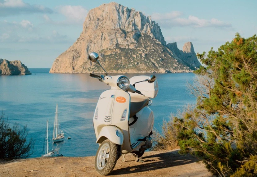 Alquiler de vespa en Ibiza con modelo frente a la isla de Es Vedrá.